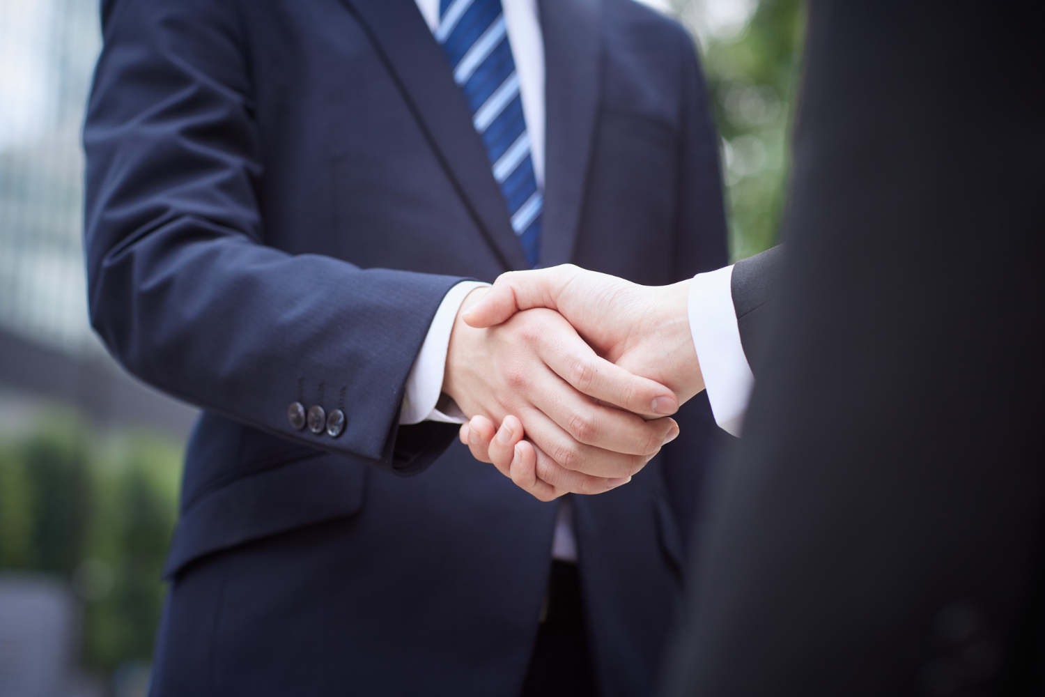 Two business people shaking hands, both in suits, outdoors.