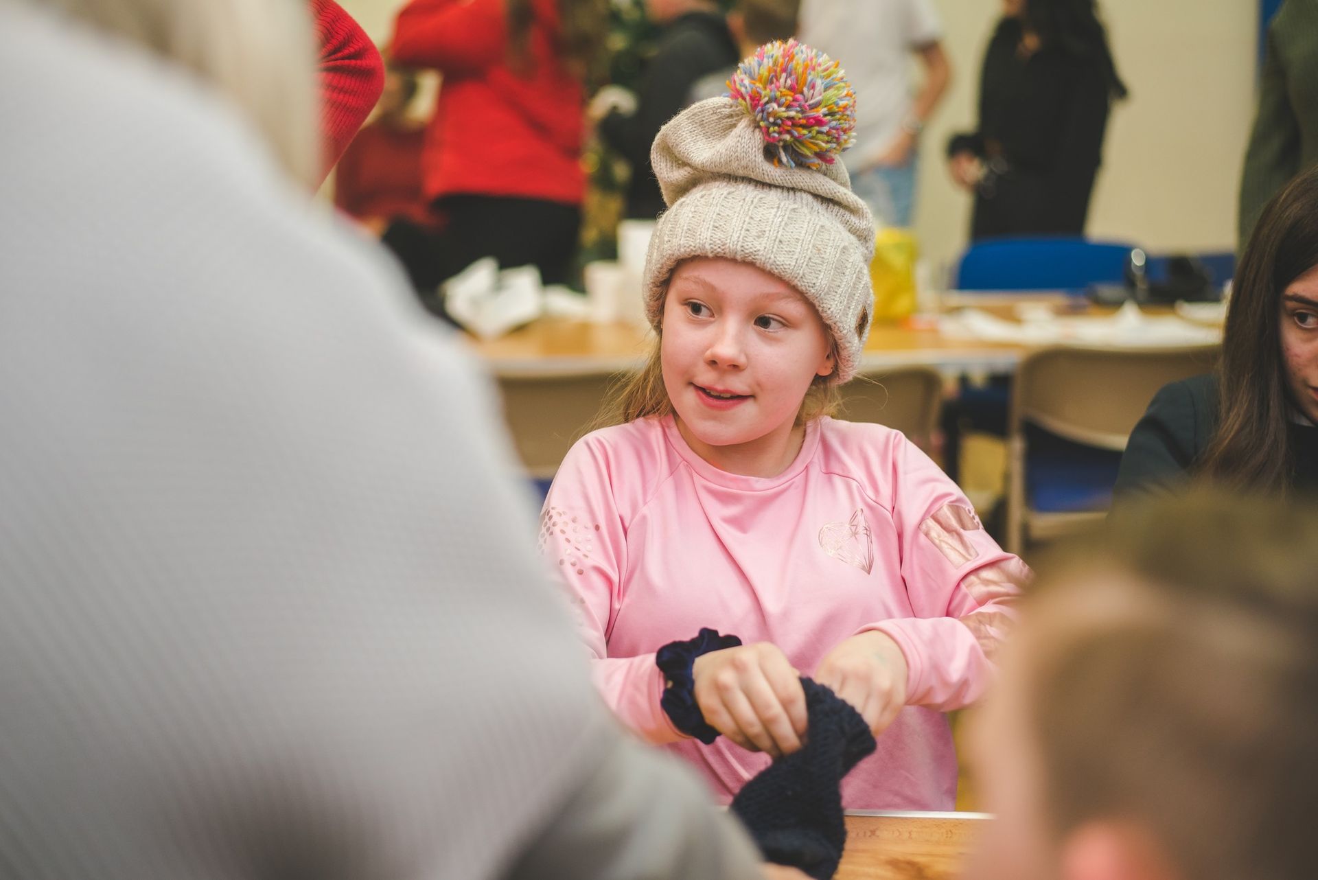 An image of a female child sat at a table wearing a pink jumper, a cream woolly hat. She is putting on a pair of black gloves and looking into the distance.