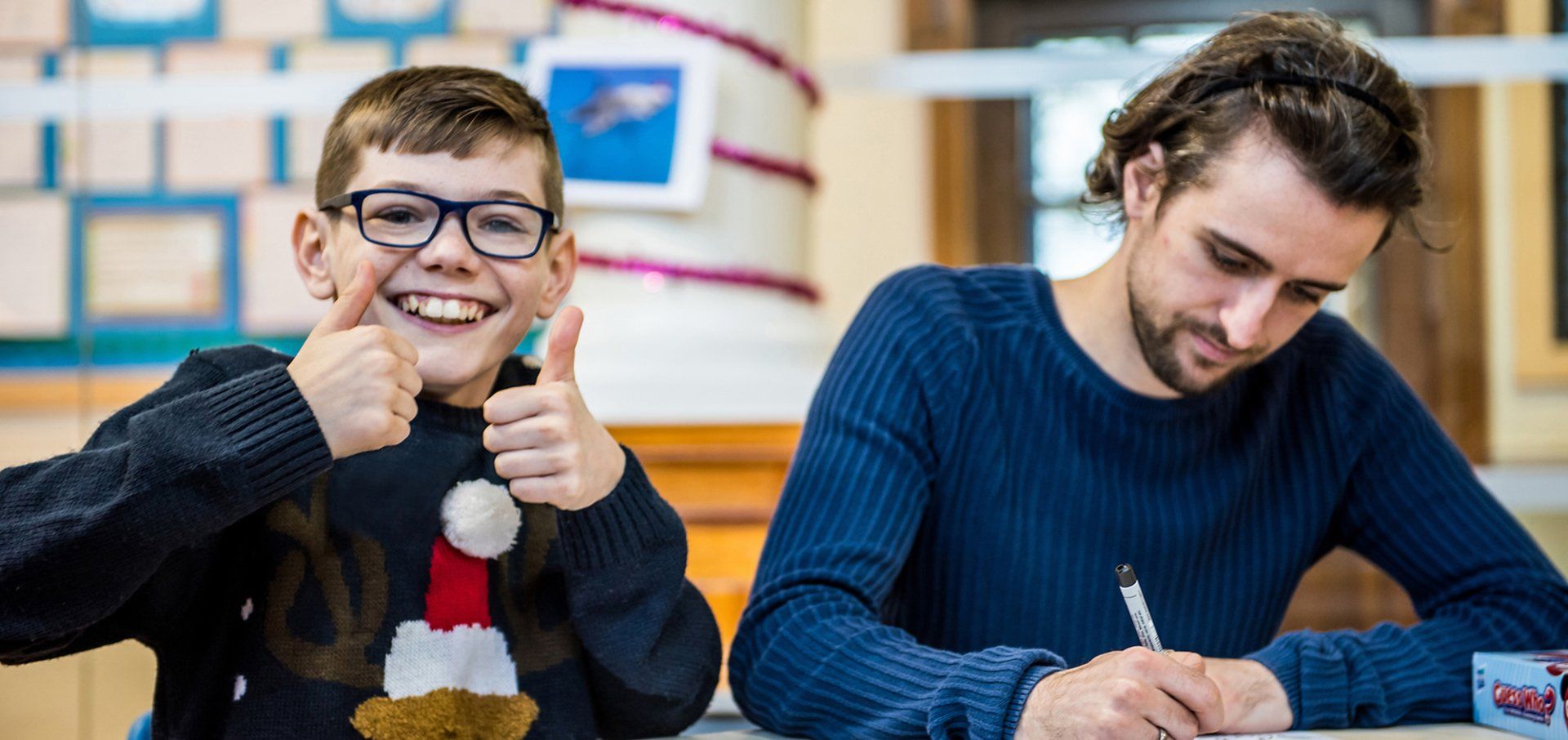 An image of an male child and an adult man sat on a sat. The child is wearing a festive jumper and is smiling directly at the camera with his thumbs up. The adult is wearing a blue jump and is writing on a piece of paper