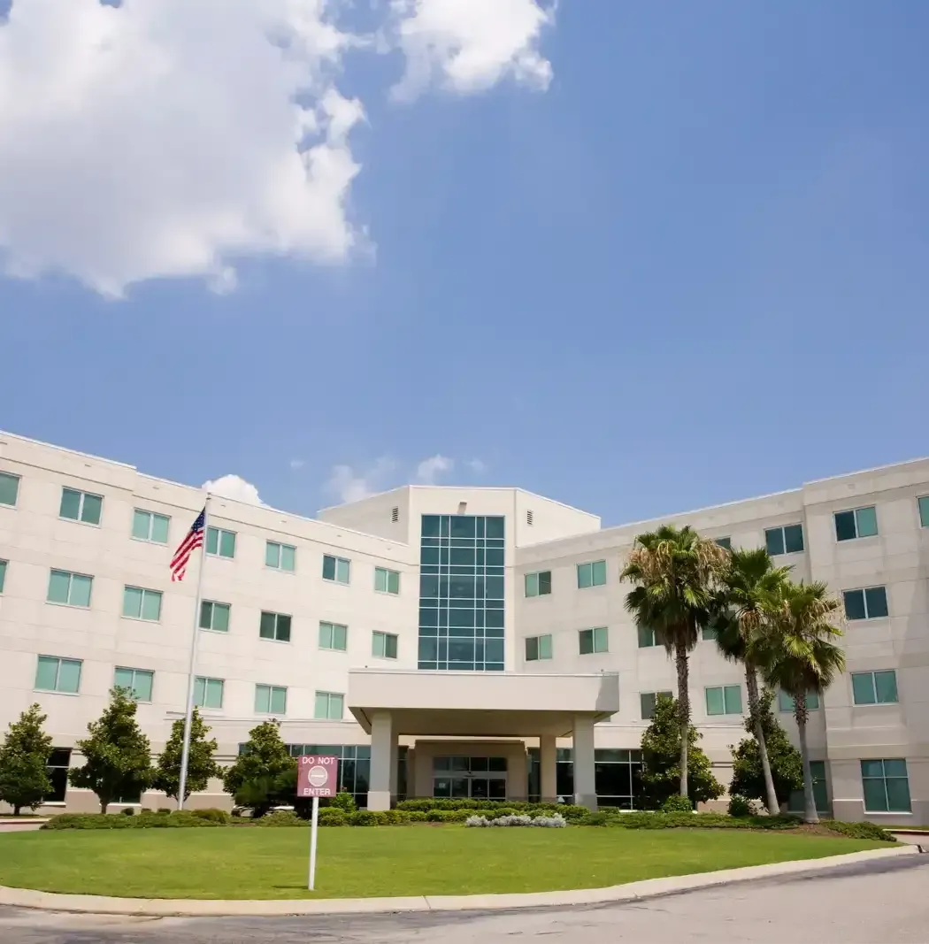 White multi-story building with a central entrance, palm trees, and a U.S. flag under a blue sky