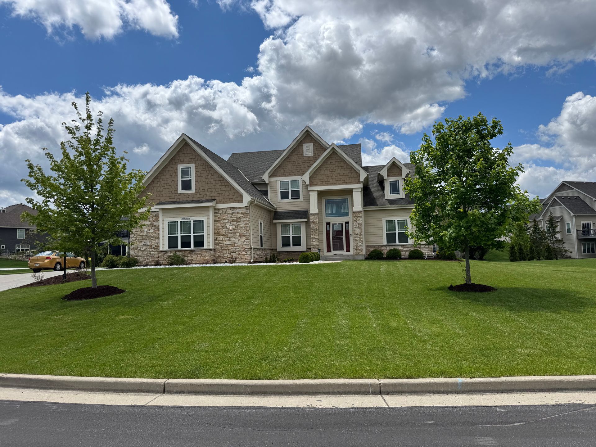 Large two-story house with tan brick and stone exterior. Green lawn, two young trees, blue sky with clouds.