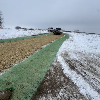 Two trucks driving on a gravel road covered with snow; a green erosion control blanket is visible.
