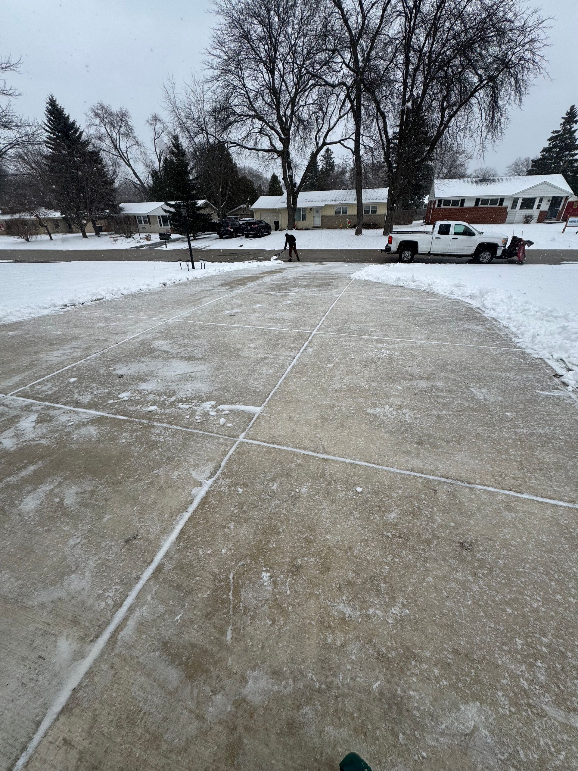 Snowy driveway with a person shoveling and a truck plowing snow. Houses and trees in the background.