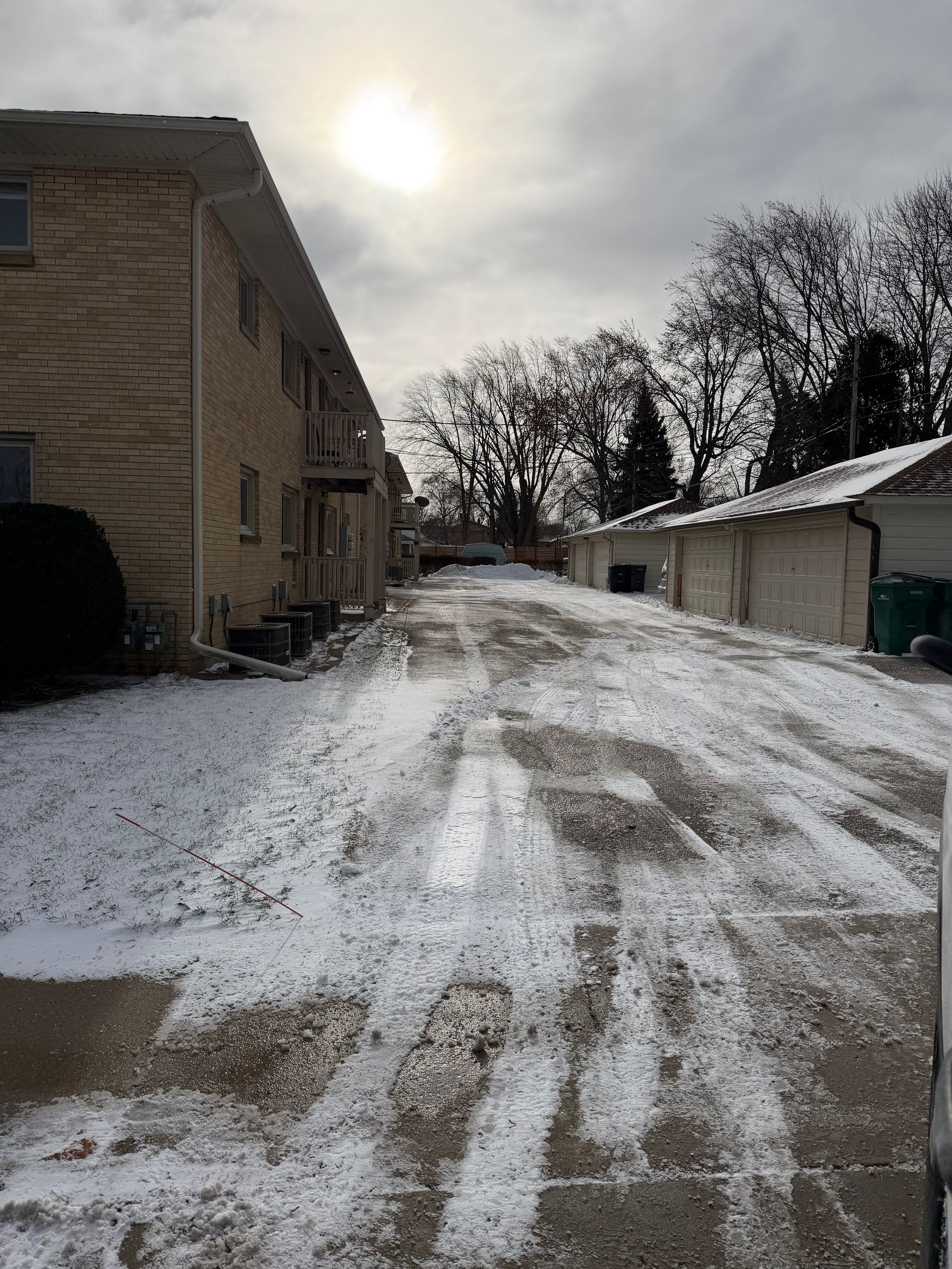 Snow-covered driveway between a two-story apartment building and garages under a cloudy sky.