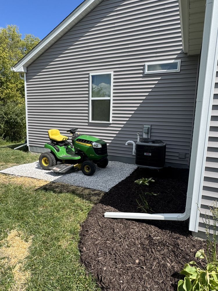 Green John Deere riding lawnmower parked on gravel next to a house with gray siding.