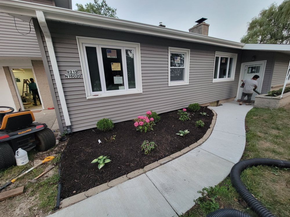A house with gray siding, white window frames, and a concrete path. Workers are visible, and there is landscaping in front.