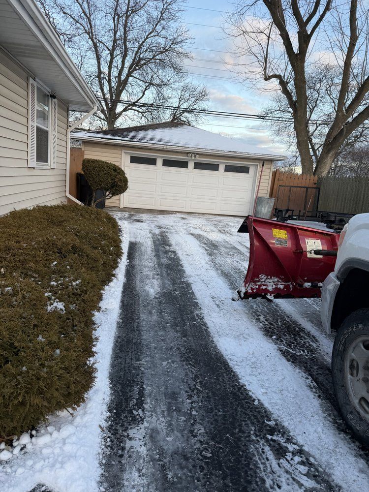 Snowy driveway with a plow attached to a truck, leading to a garage. A house is on the left.