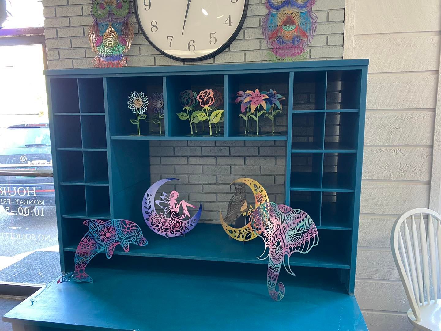 Teal cabinet with shelves displaying colorful metal art: flowers, crescent moons with figures, and an elephant.