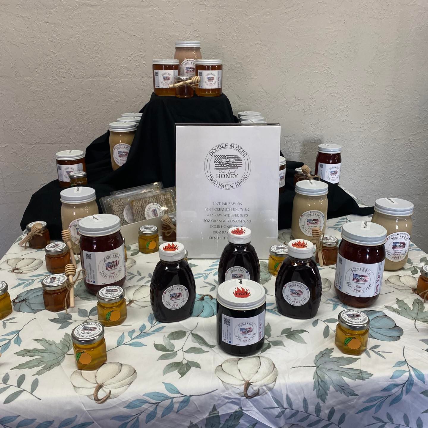Jars of preserves and honey on a table covered with a leaf-patterned tablecloth. A paper bag is centered.