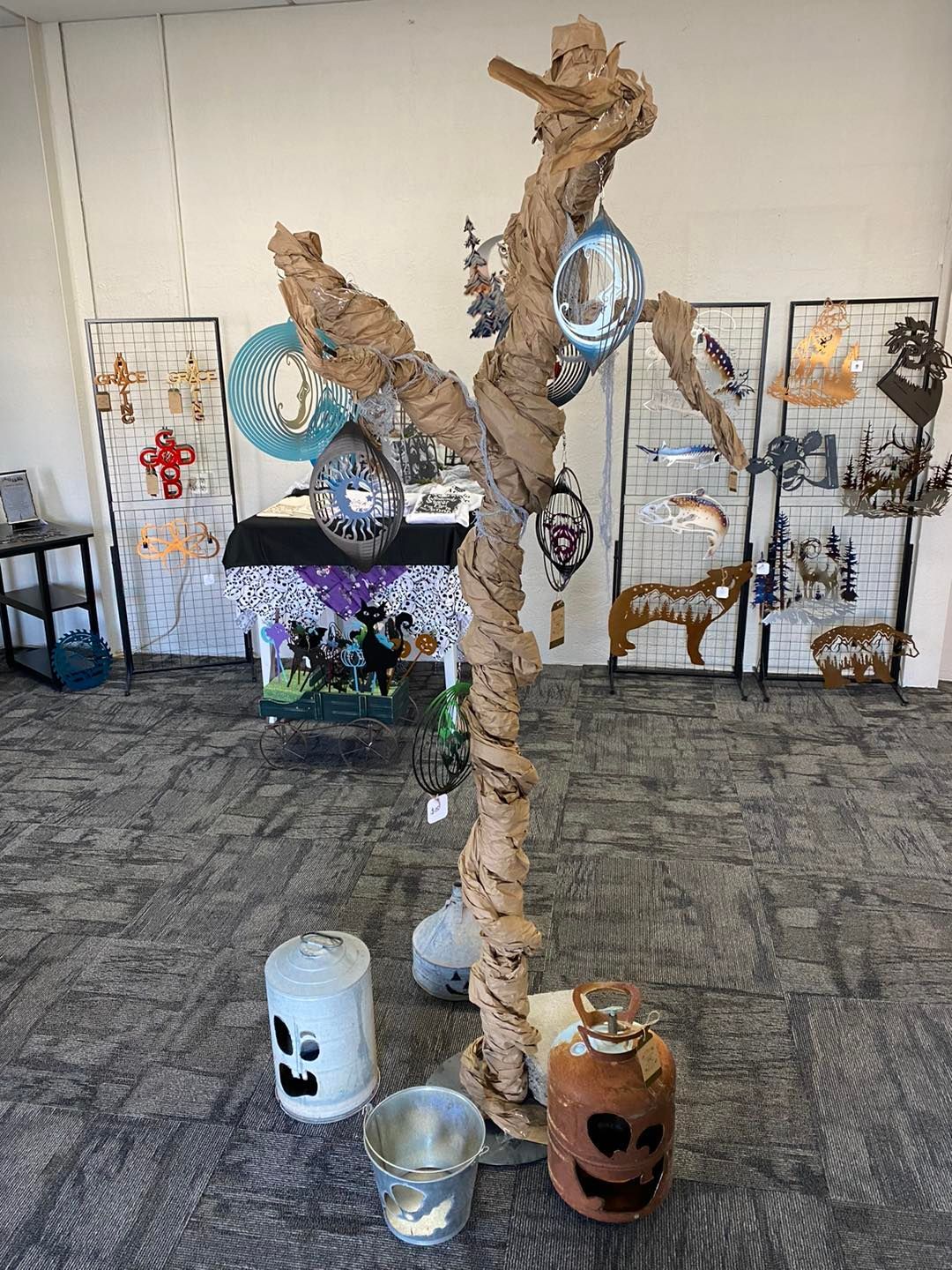 A tall, twisted tree sculpture wrapped in brown paper, with hanging ornaments and pumpkins at its base.