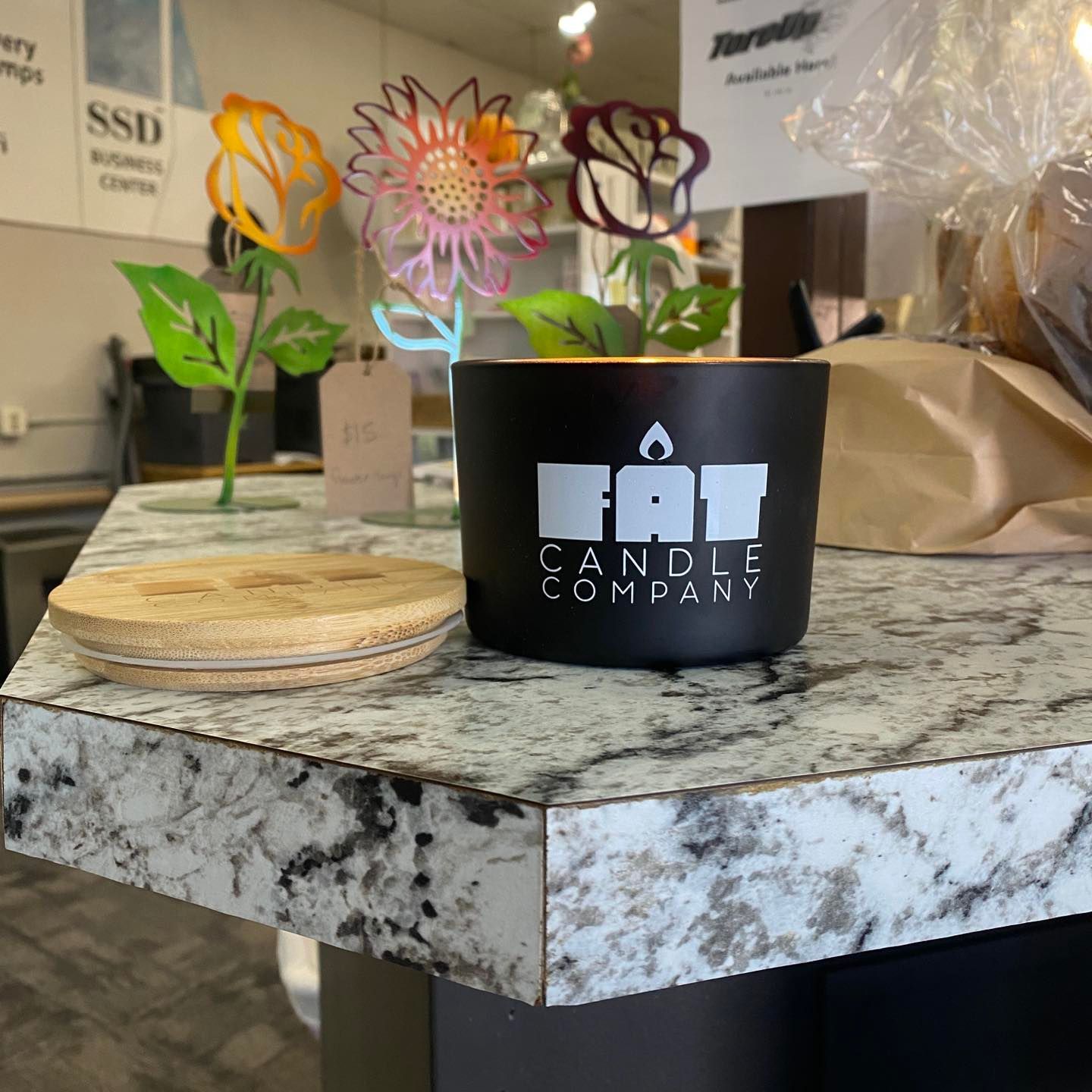 Black Fat Candle Company candle on a countertop with a cork coaster and metal flower art in the background.