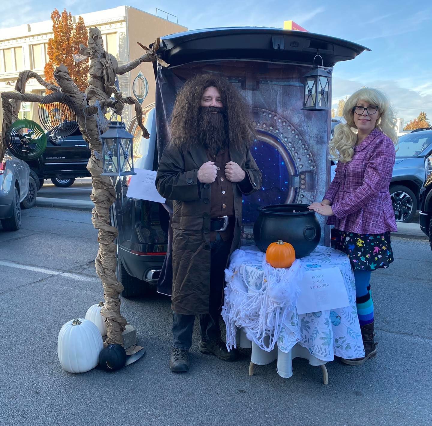 Two people in Harry Potter costumes at a decorated car trunk. Includes a cauldron, pumpkins, and a tree prop.