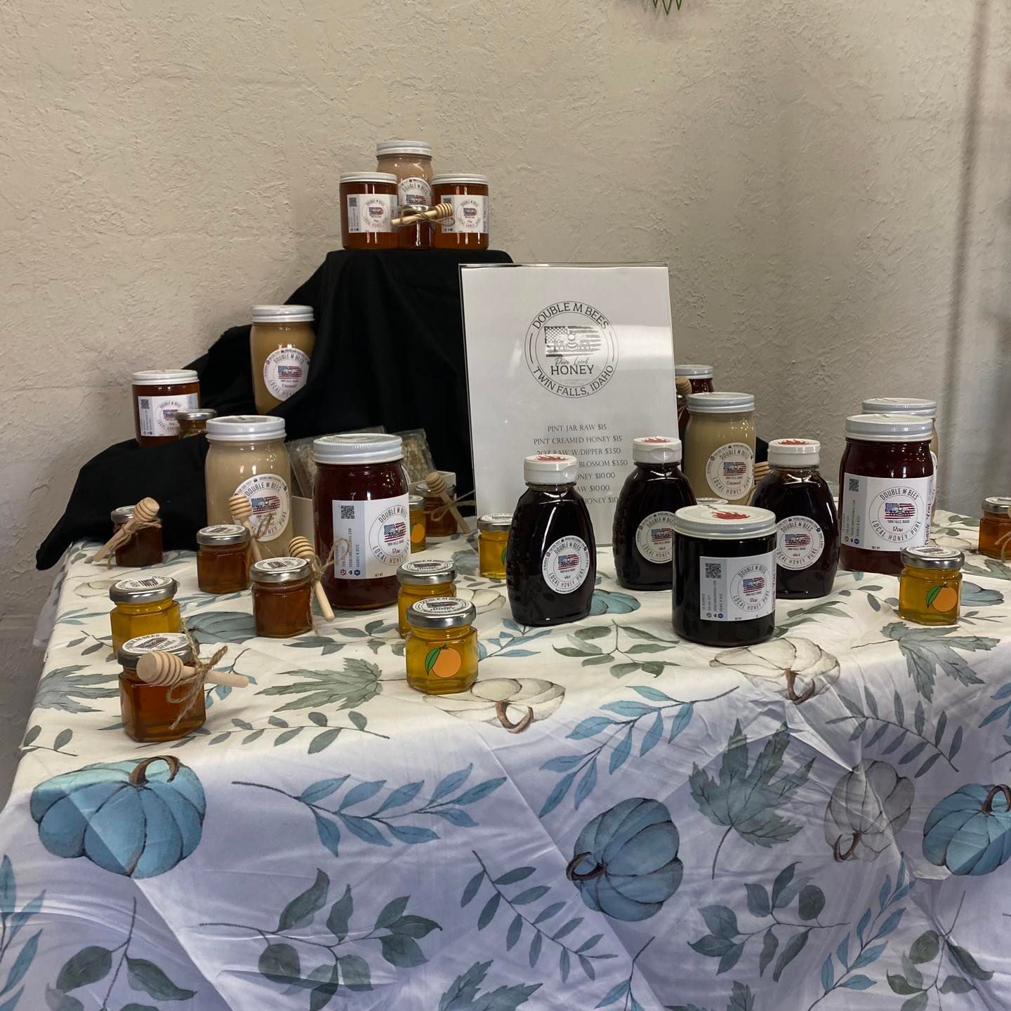 Table of honey jars for sale, varying colors and sizes, with a sign. White table cloth with blue pumpkin pattern.