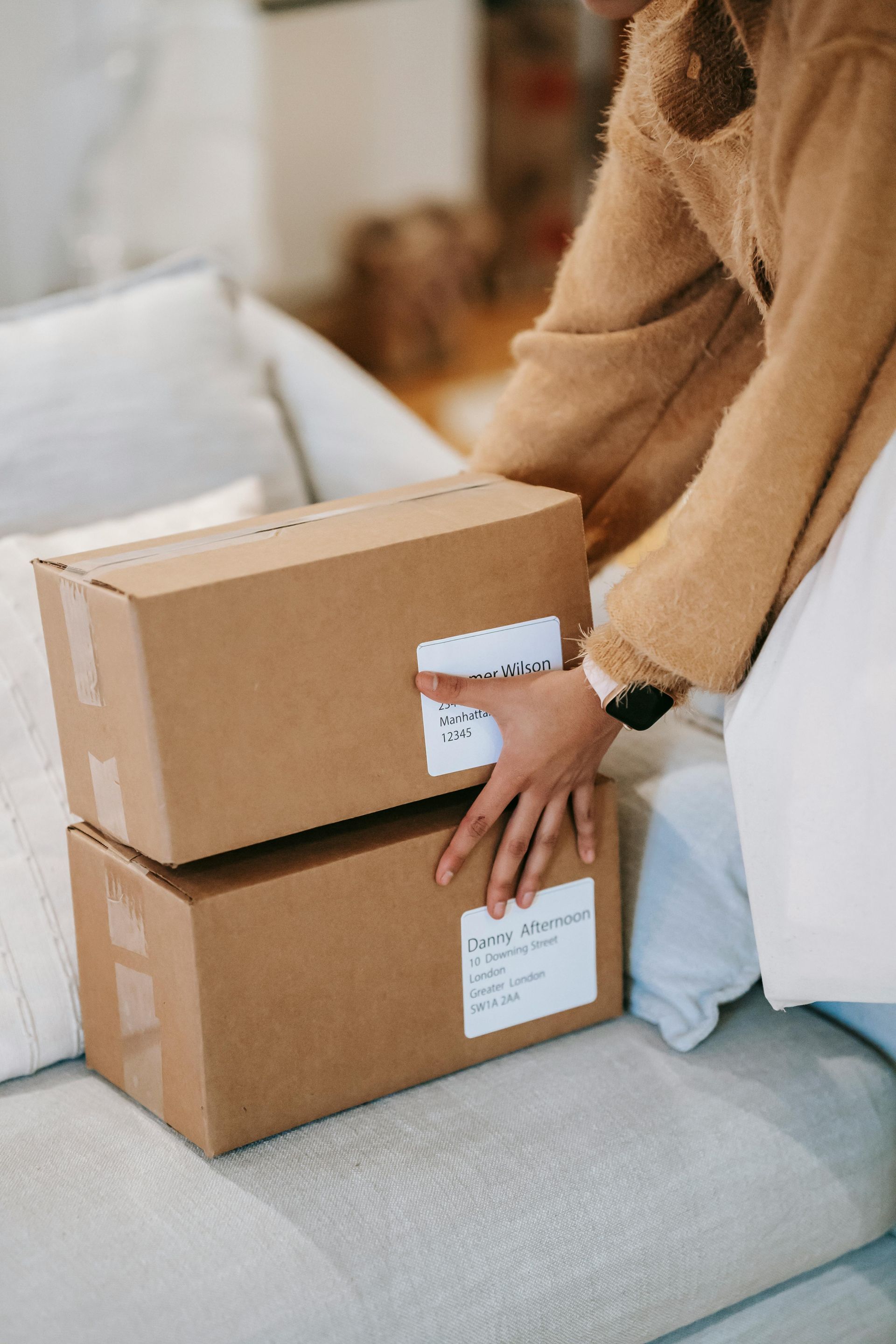 Person on couch holding two stacked cardboard boxes with shipping labels.