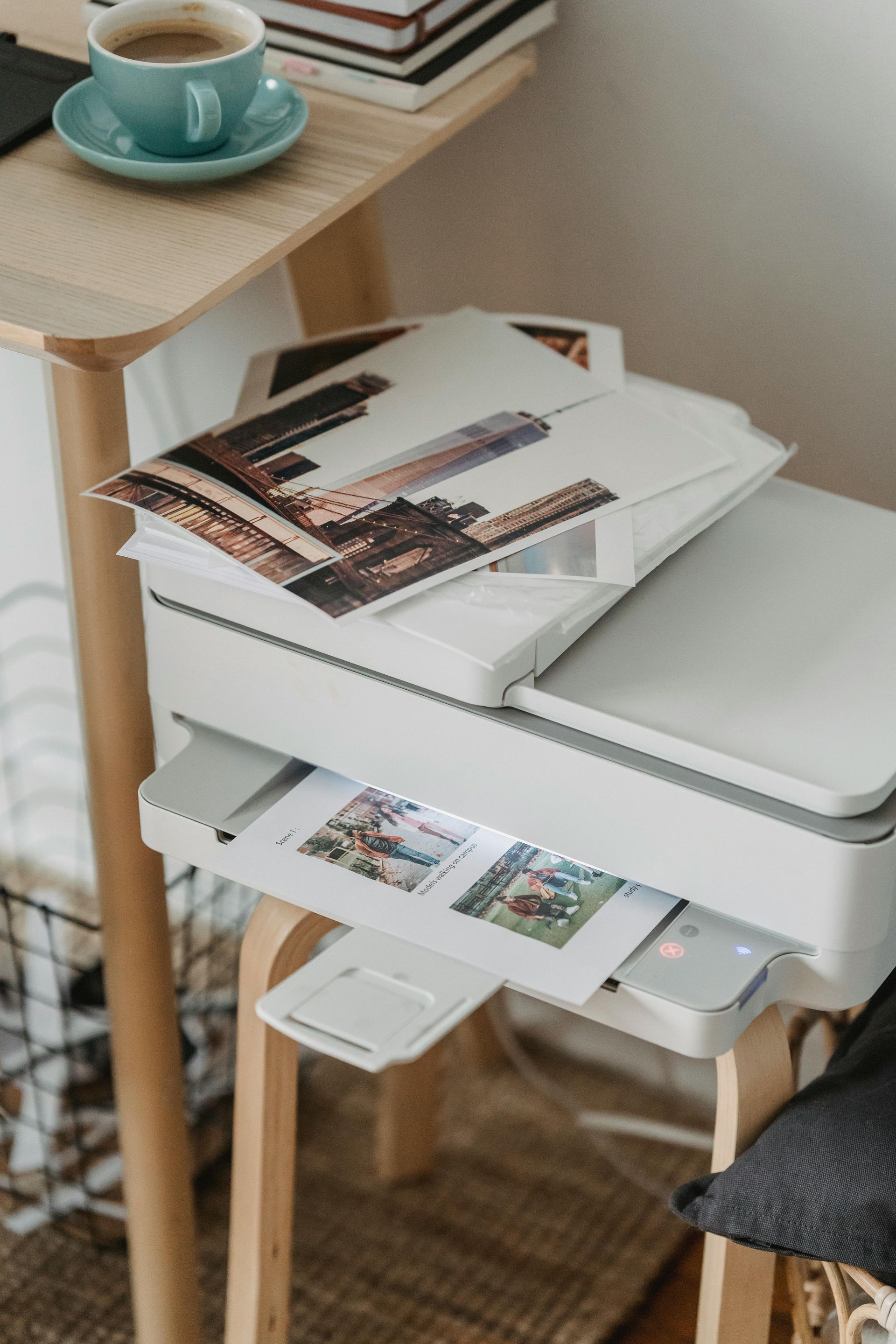 White printer on a wooden stool with printed photos. A mug of coffee sits on a small desk beside it.