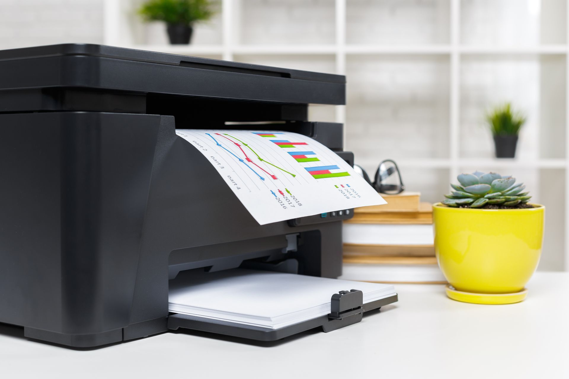 Black printer printing a document with colorful charts on a white desk with a yellow pot of succulent.