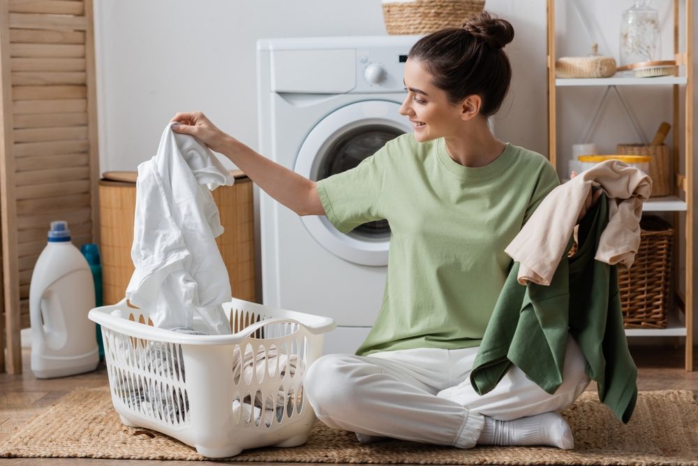 Happy young woman sorting clothes near washing machine in laundry room