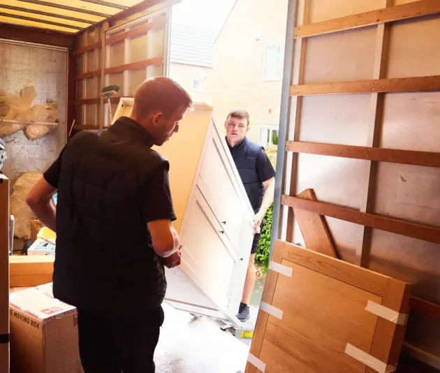 Two movers loading furniture into a truck. One holds a large panel. Boxes and other items are visible.