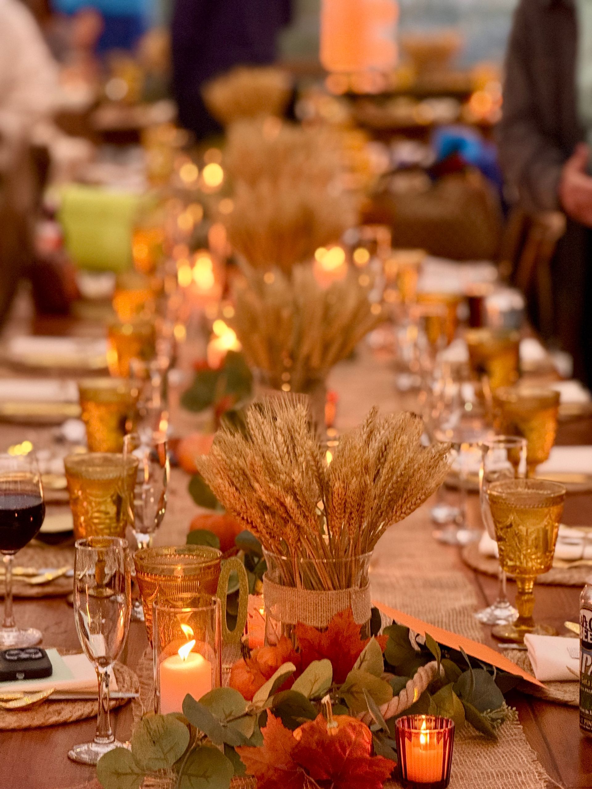 Long dining table set for a fall celebration with wheat stalks, candles, and colorful leaves.