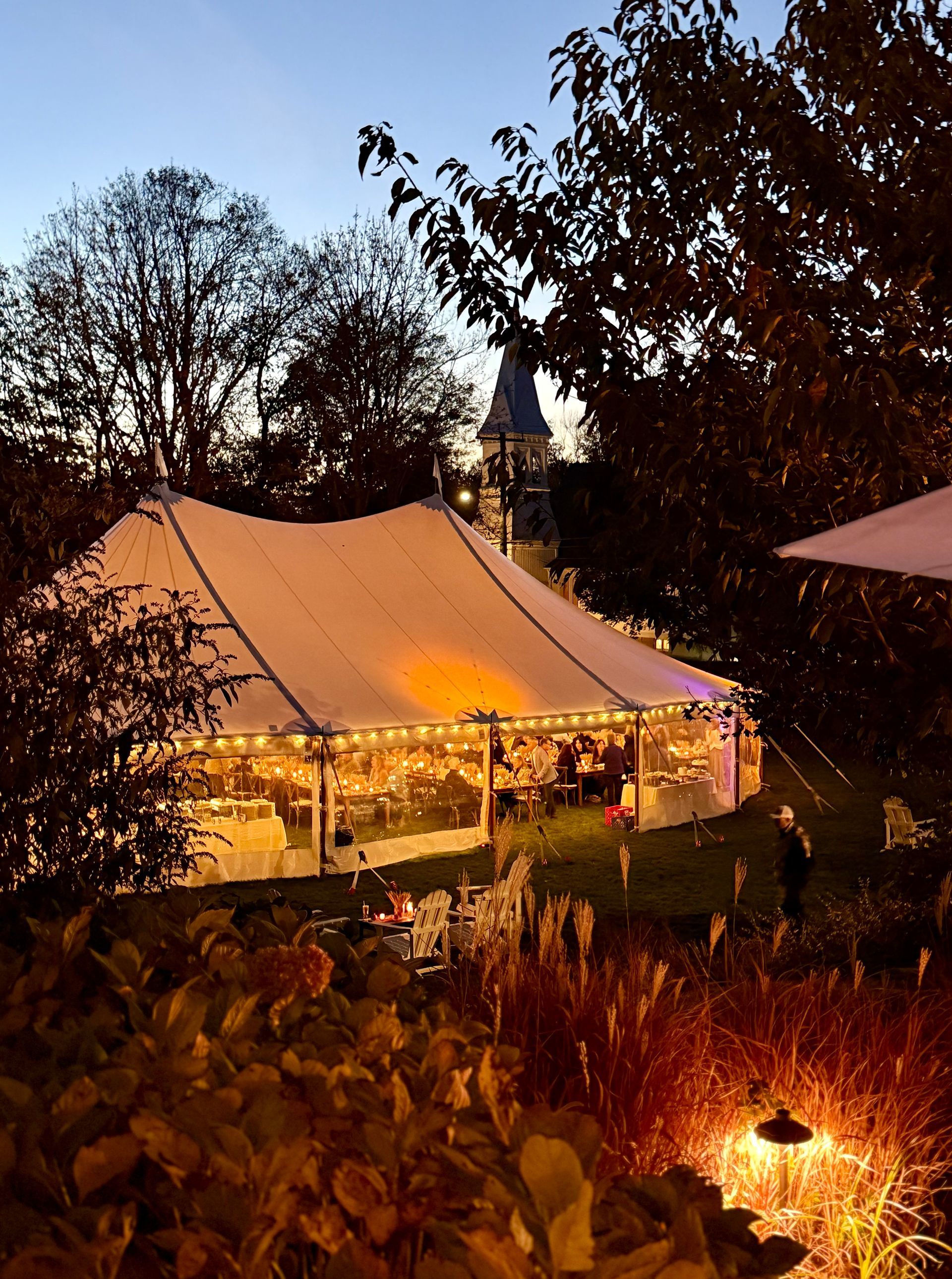 A white tent illuminated at dusk, set on a lawn, with a church steeple in the background.