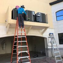 A man is standing on a ladder fixing air conditioners on the side of a building.