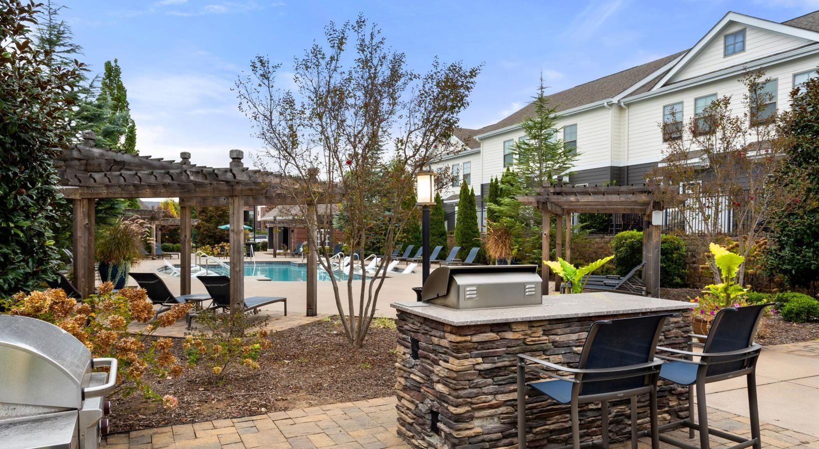 A patio area with a grill and chairs in front of a swimming pool.