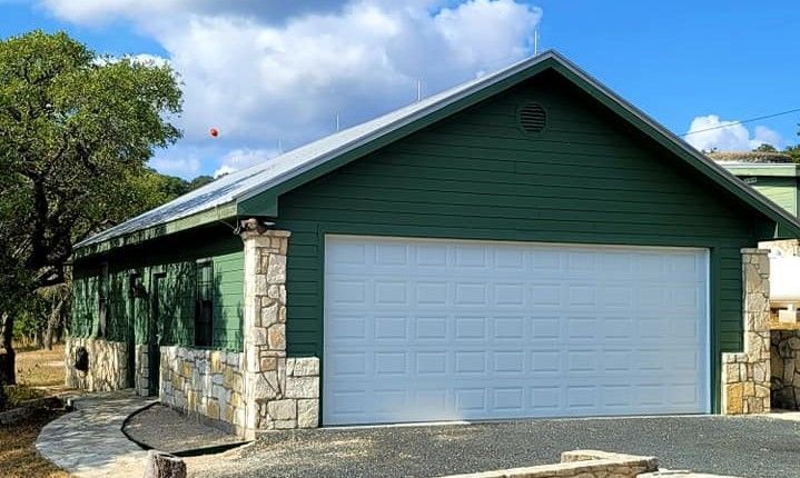 Green garage with stone siding and white doors under a blue sky, viewed from the driveway