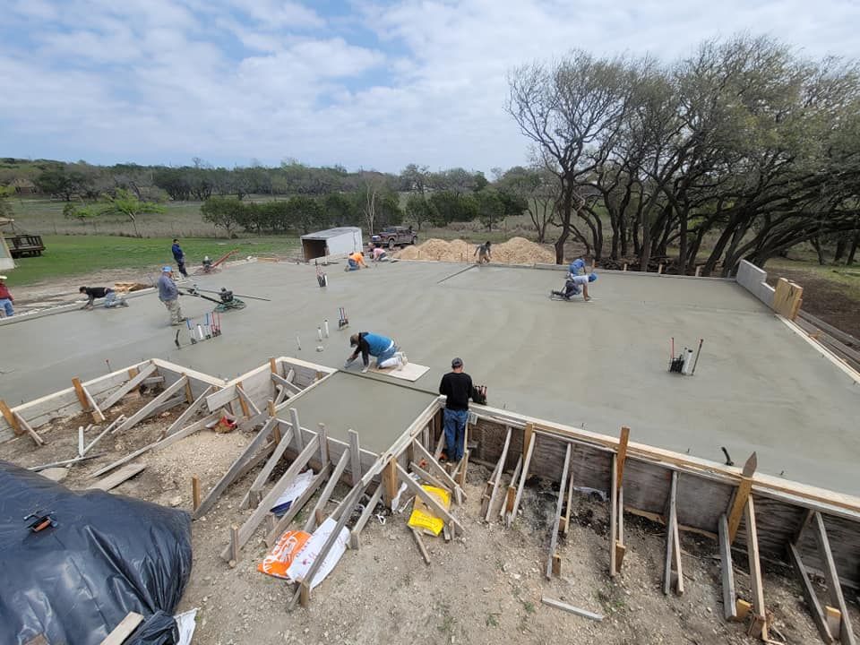 Construction workers pouring a concrete foundation on a large outdoor site with wooden forms and trees nearby