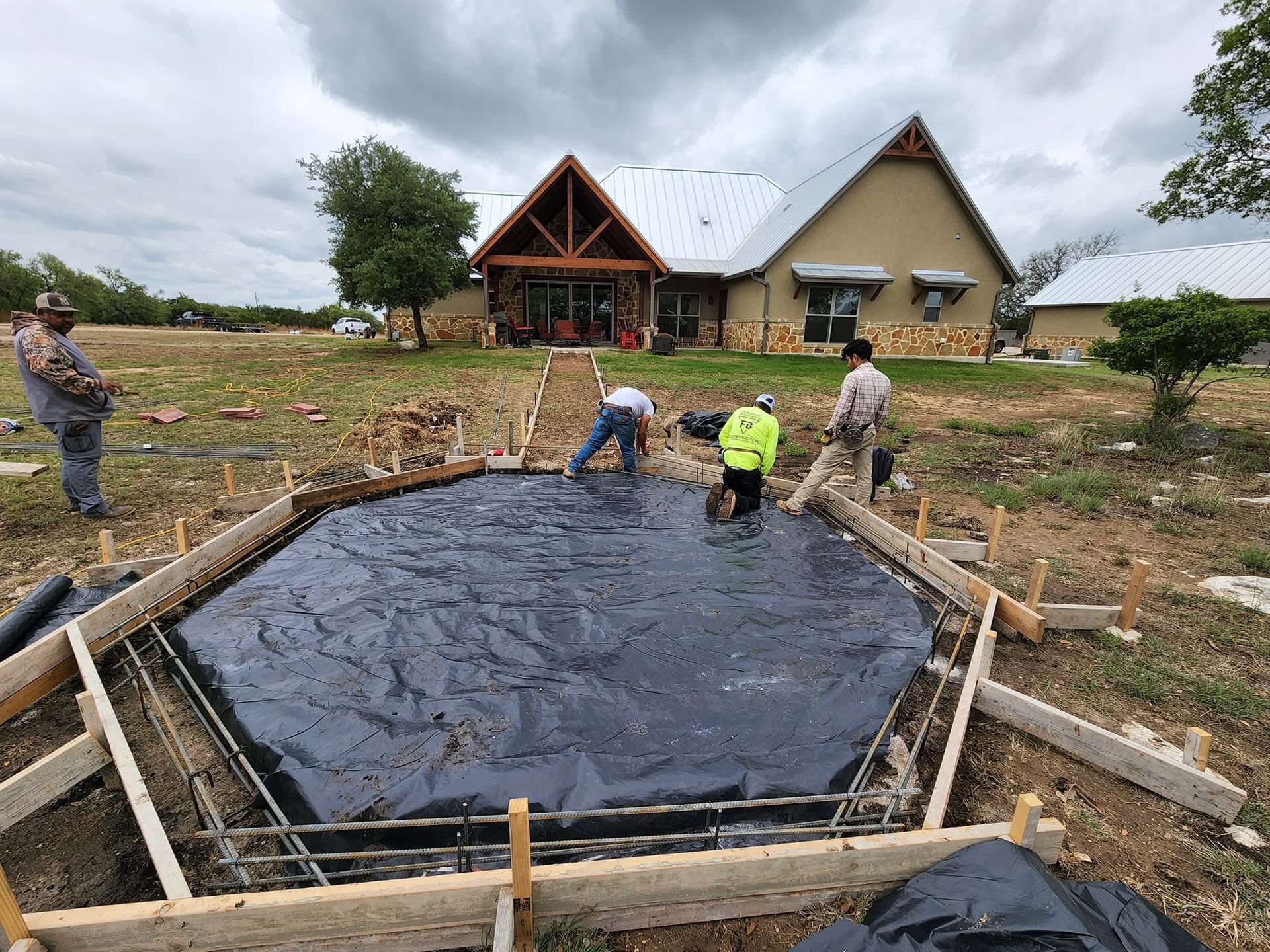 Workers lining a tarp-lined pond foundation at a rural construction site with two houses in the background