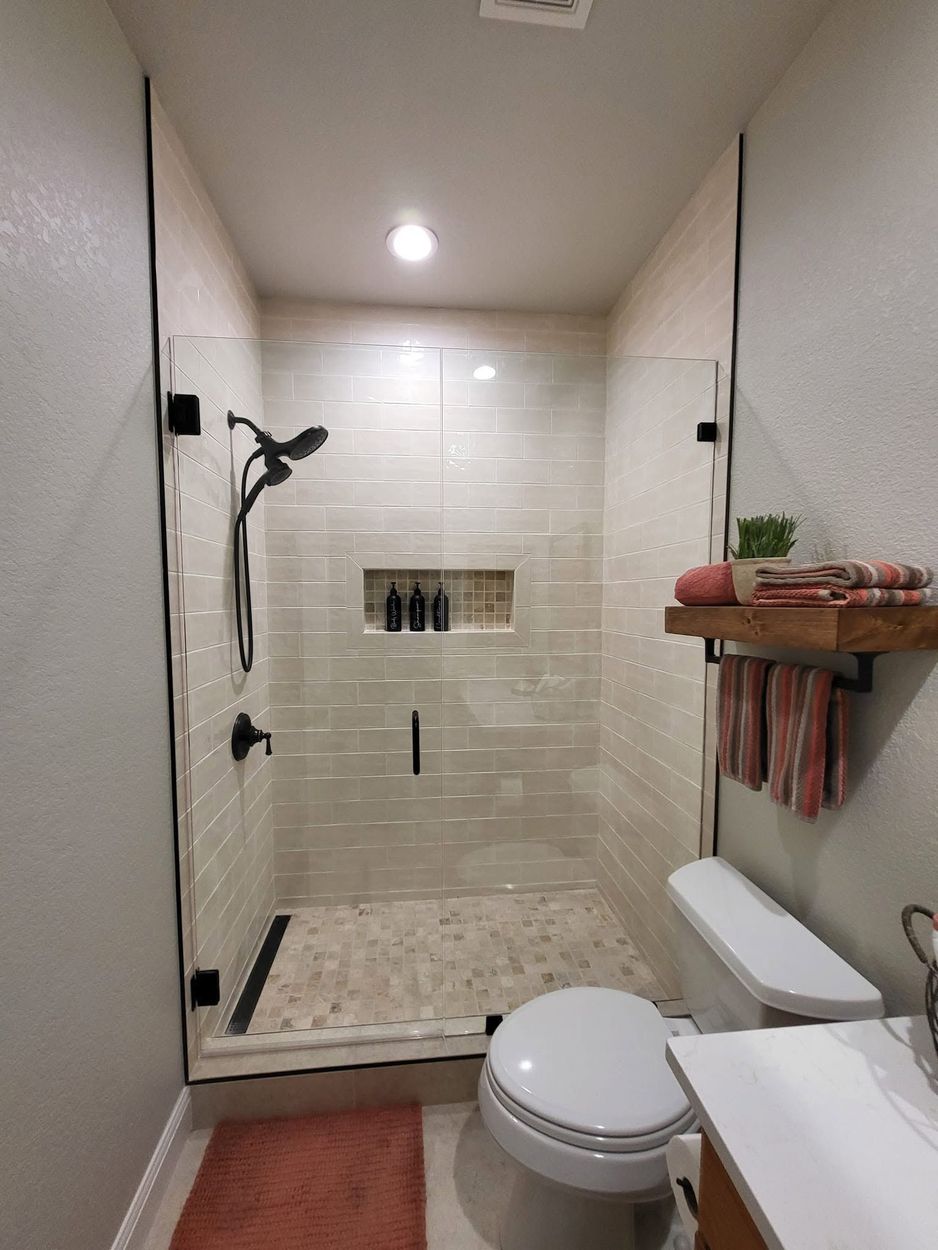Small tiled walk-in shower with white walls, shower head, built-in shelf, and toilet in the foreground.