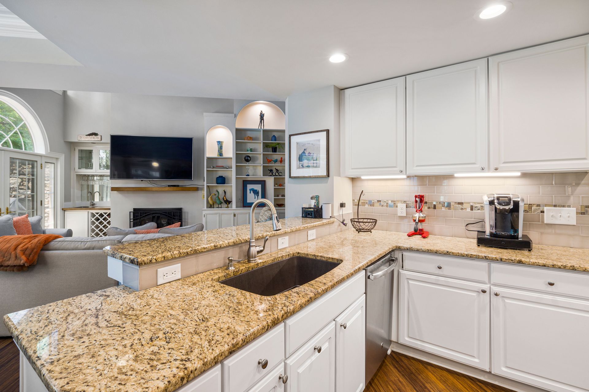 Bright kitchen with white cabinets, granite island, and open view to a living room with fireplace and TV