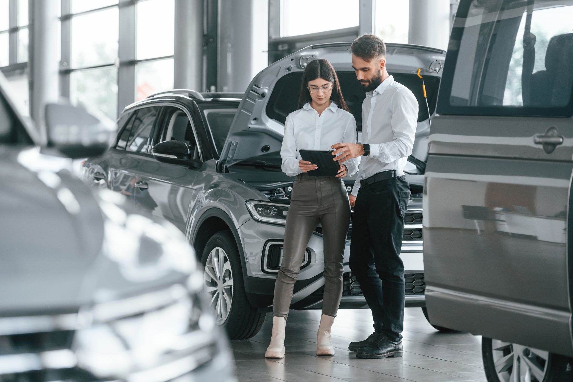 Mulher e homem revisando documentos em uma concessionária de carros, SUVs cinzas ao fundo.
