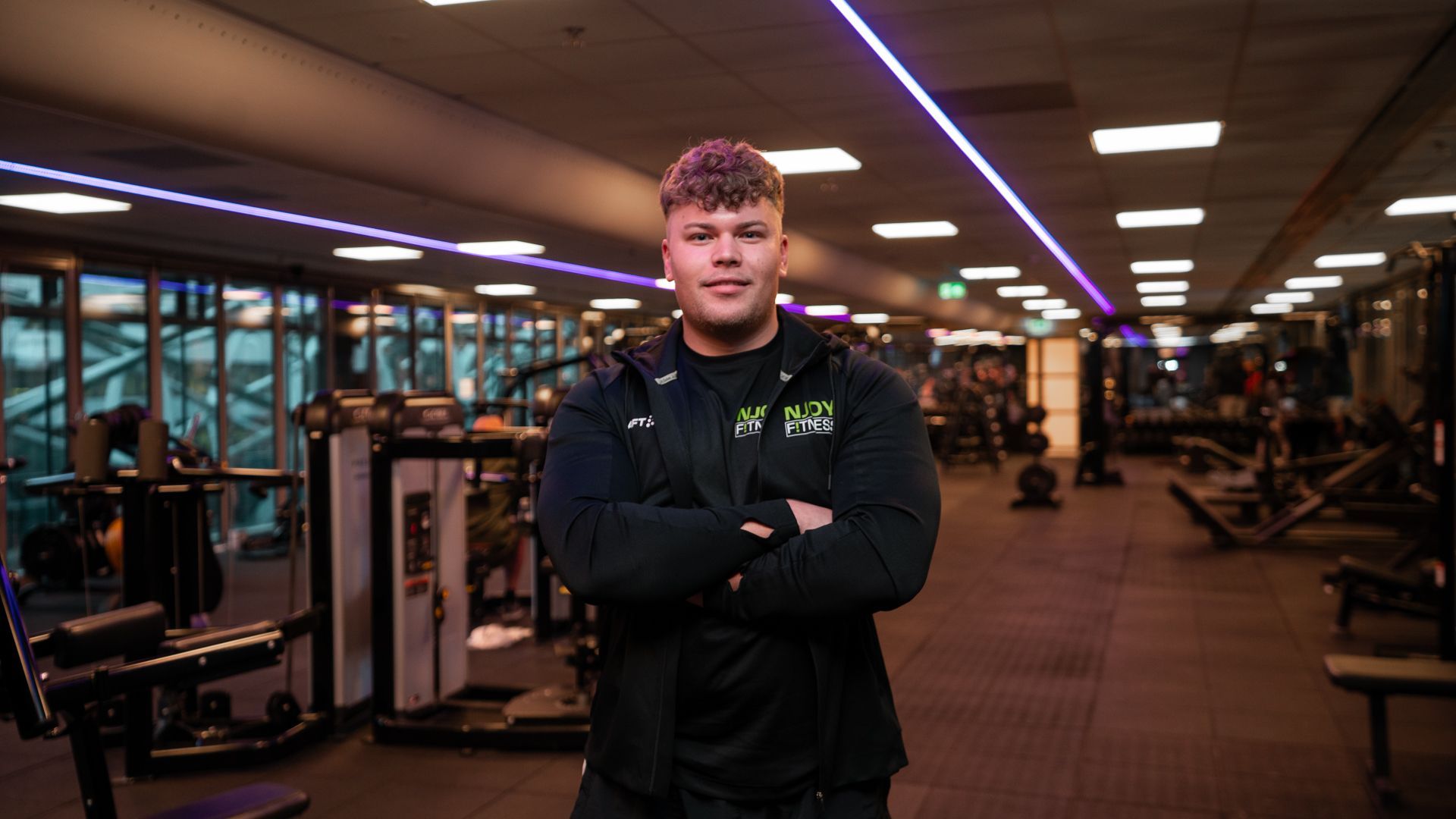 Een man in een sportschool, met de armen over elkaar, draagt ​​een zwarte jas en poseert voor fitnessapparaten.