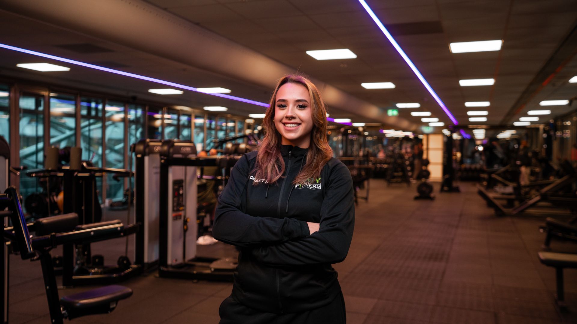 Een vrouw in zwarte sportkleding lacht met haar armen over elkaar in een sportschool.