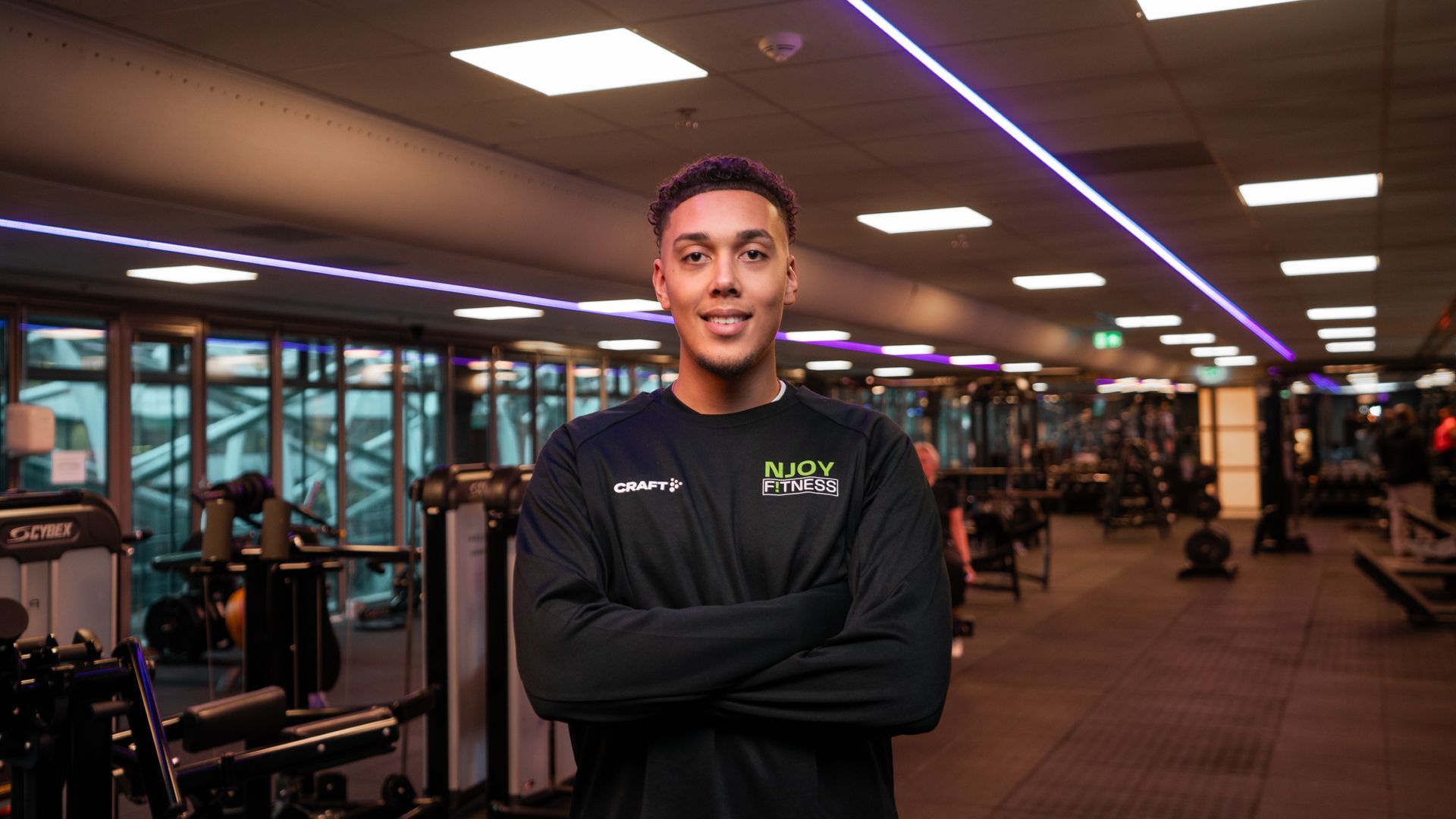 Een man in sportkleding met de armen over elkaar, lachend in een sportschoolomgeving.