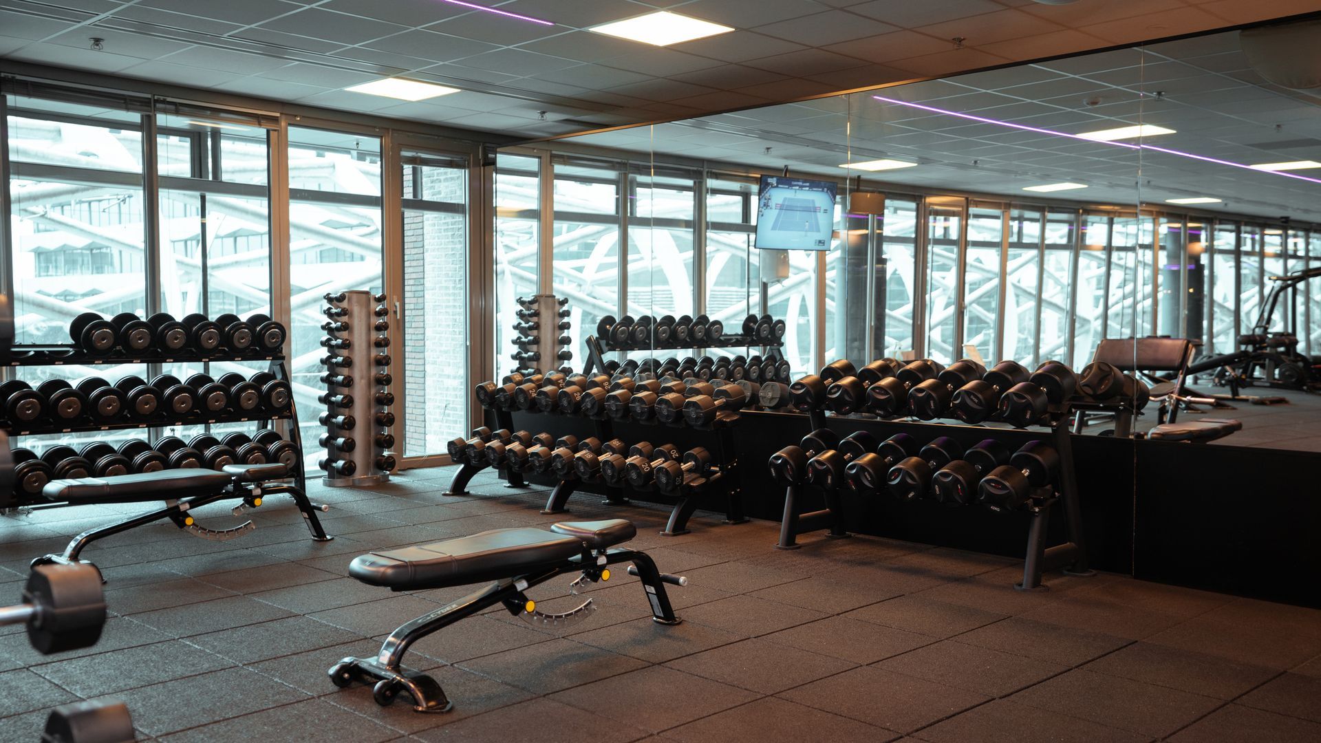 Interieur van een sportschool met halterrekken, een gewichtsbank en grote ramen.