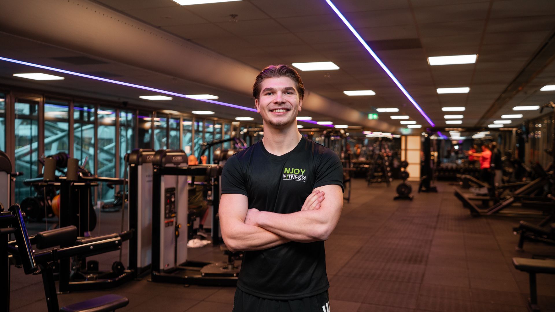 Man met gekruiste armen lachend in een sportschool. Donker shirt, apparatuur op de achtergrond, fel licht.