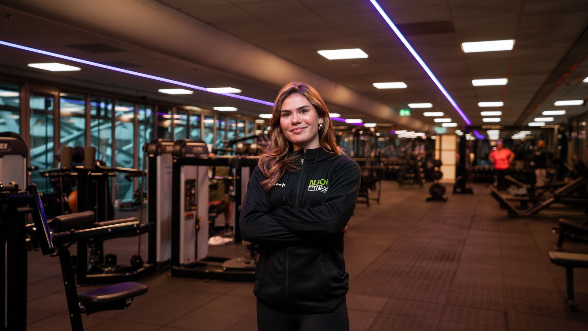 Vrouw in de sportschool, armen over elkaar, lachend. Draagt ​​een zwarte jas, fitnessapparatuur op de achtergrond.