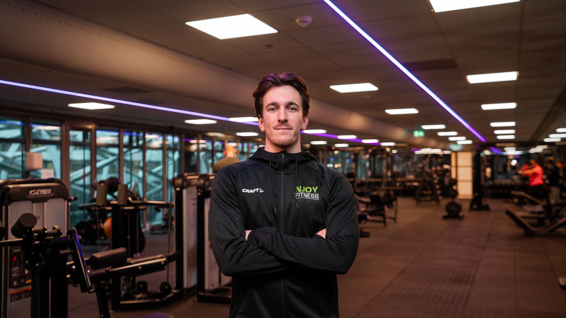 Een man in zwarte sportkleding staat met gekruiste armen in een sportschool; de apparatuur is zichtbaar.