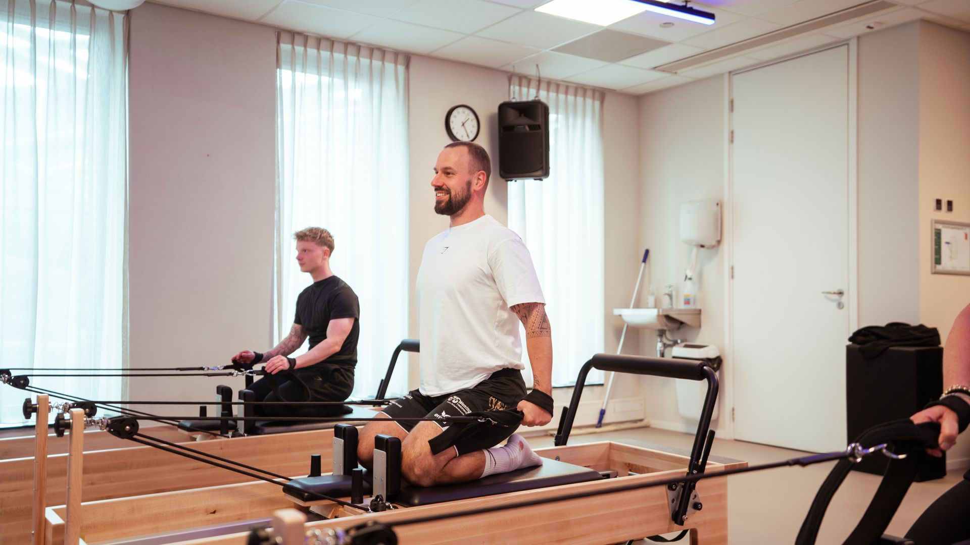 Mensen doen oefeningen op Pilates reformers in een studio. Een man staat te glimlachen, een ander knielt. Lichte omgeving.
