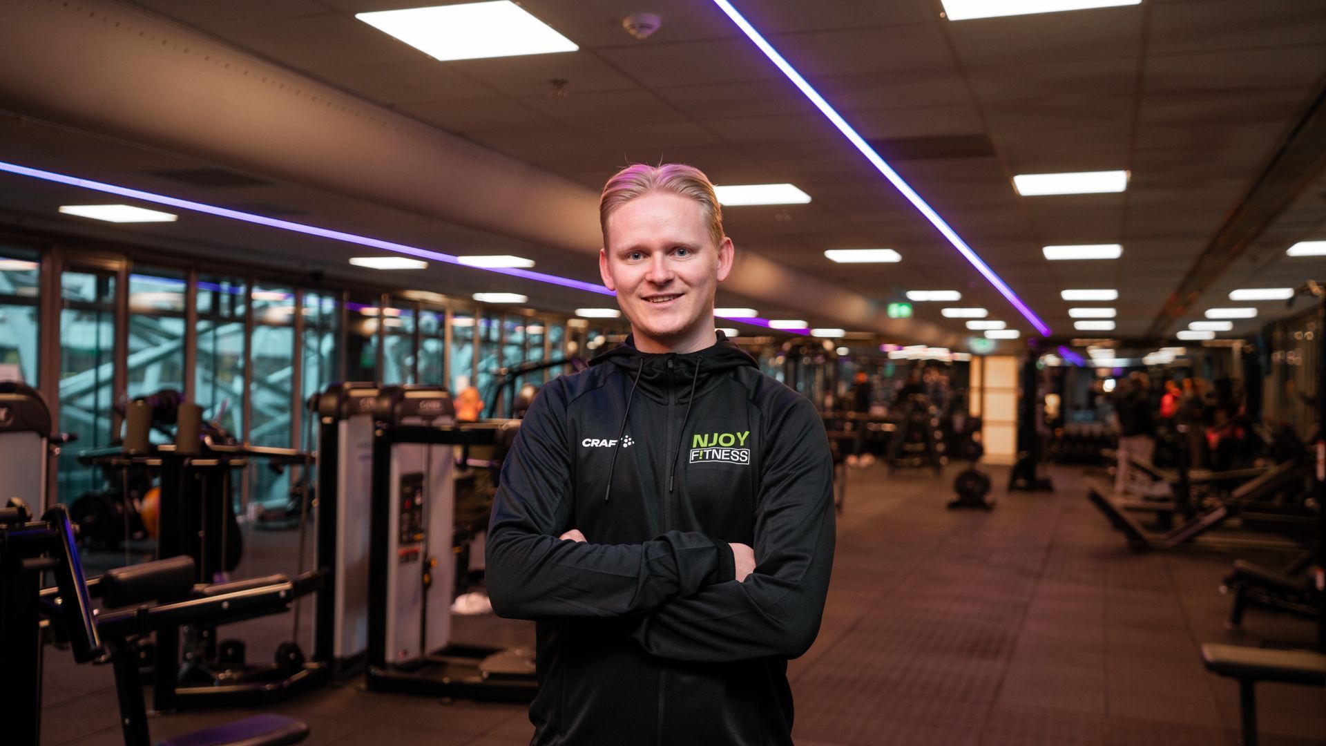 Een man in een zwarte jas met de armen over elkaar staat in een sportschool tussen de fitnessapparaten.