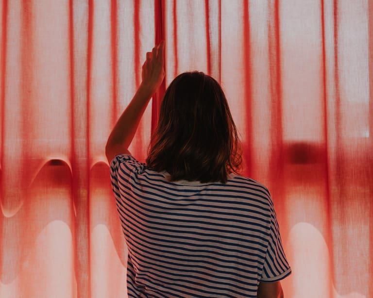 Femme en chemise rayée, vue de dos, ouvrant des rideaux rouges.