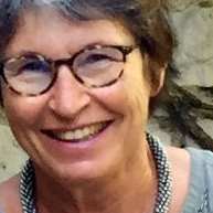 Femme à lunettes souriante, portant un collier de perles, devant un mur de pierre.