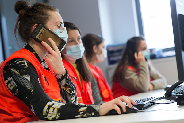 Quatre personnes portant des masques et des gilets orange à des bureaux, parlant au téléphone et utilisant des ordinateurs dans un bureau.