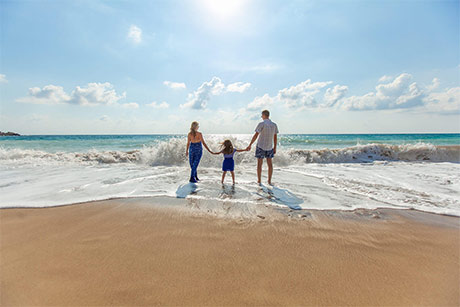 Famille de trois personnes se tenant la main, debout dans les vagues de l'océan sur une plage ensoleillée.
