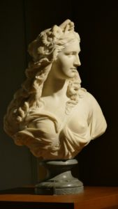 Buste en marbre d'une femme aux cheveux flottants et aux vêtements drapés, posé sur un piédestal.