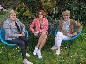 Trois femmes assises sur des chaises bleu sarcelle à l'extérieur. L'une porte un blazer gris, une autre un rose et la troisième une veste marron, souriantes.