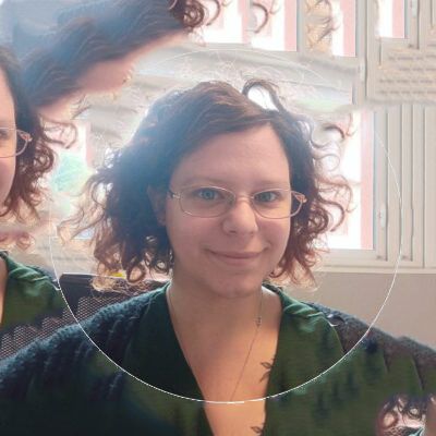 Femme aux cheveux roux bouclés et aux lunettes, souriante, vêtue de vert, à l'intérieur.