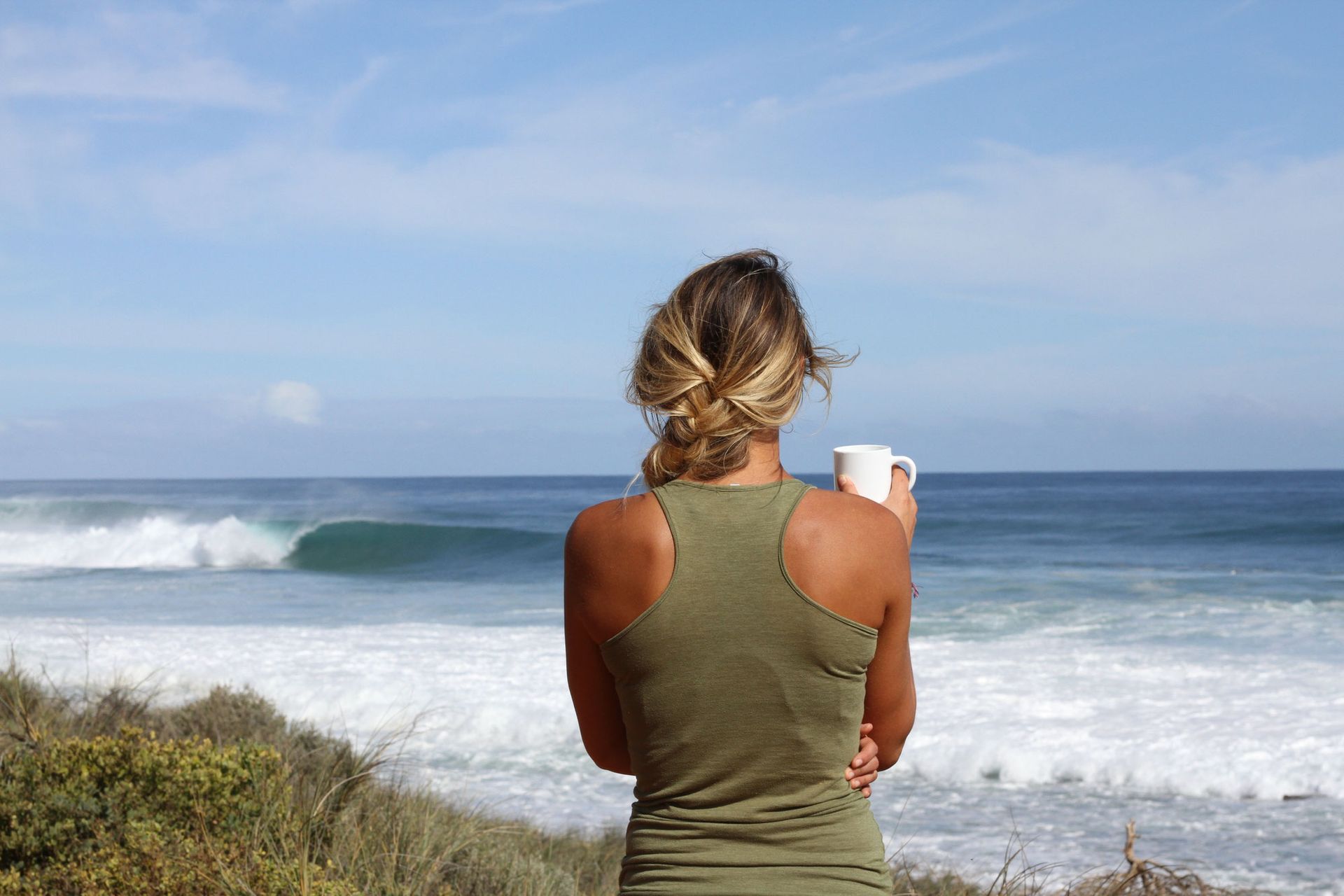 Une femme en débardeur olive tenant une tasse, regardant les vagues de l'océan depuis une falaise par une journée ensoleillée.