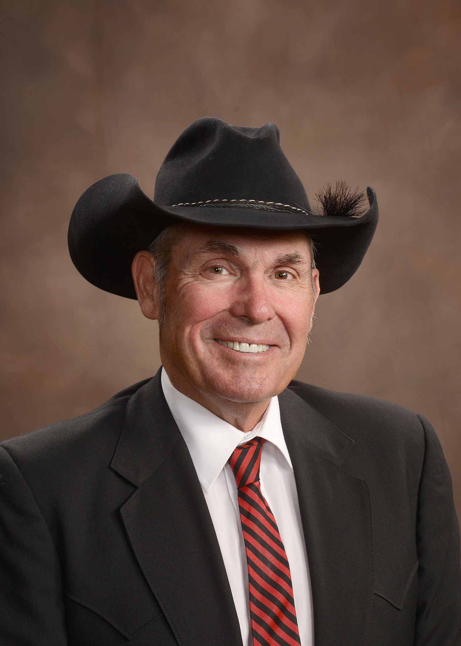 A man wearing a cowboy hat and glasses is standing in front of a brown background.